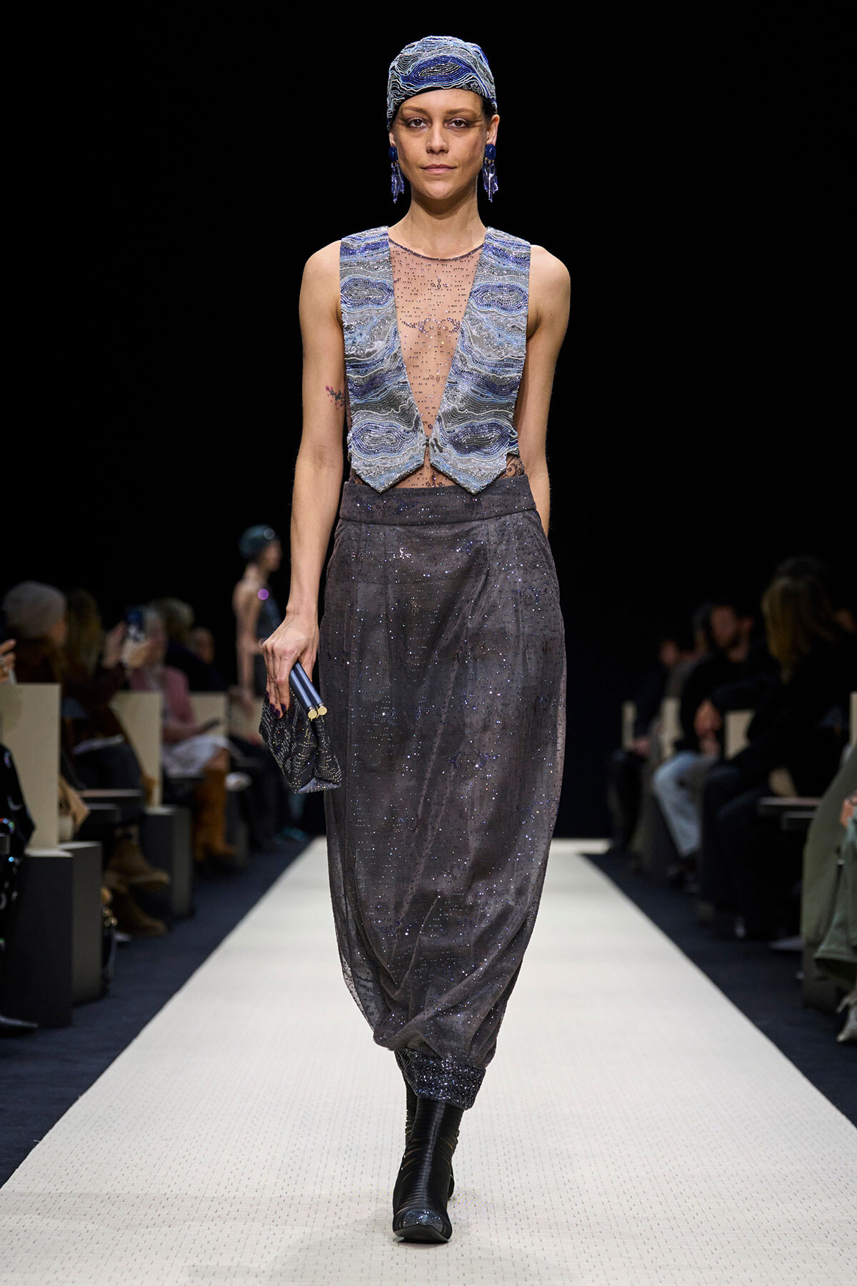 Model walks a white runway in a metallic blue-gray ensemble: sleeveless jeweled mesh top with matching vest panels, wide harem-style sequin pants, a matching head wrap, blue earrings, and a small clutch purse in hand.