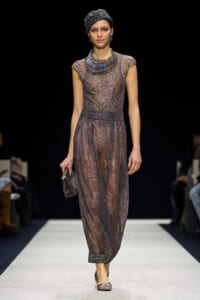 Model walking a runway in a sheer, sequined taupe gown with a structured metallic collar and matching head wrap, holding a small clutch.