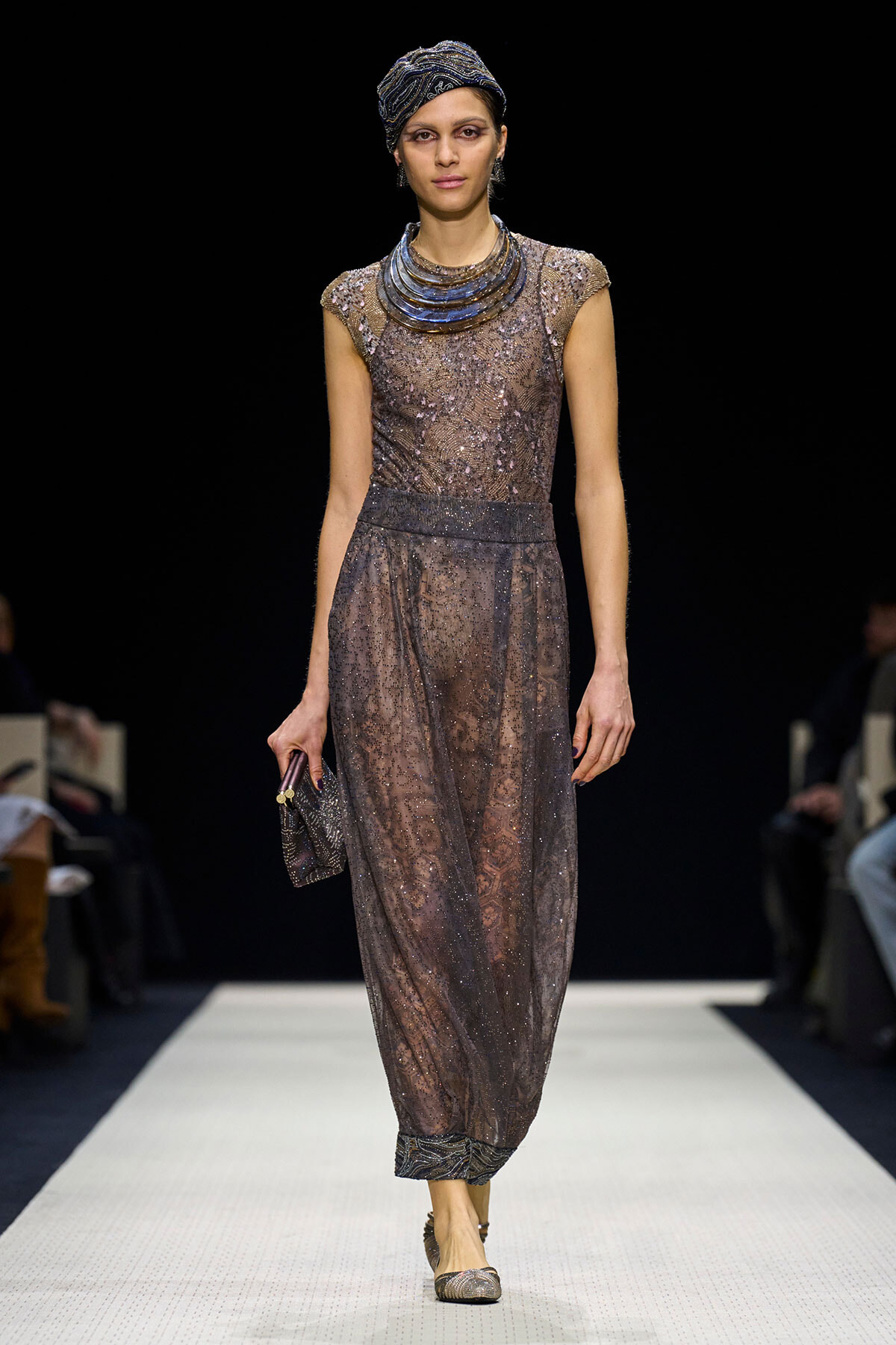 Model walking a runway in a sheer, sequined taupe gown with a structured metallic collar and matching head wrap, holding a small clutch.