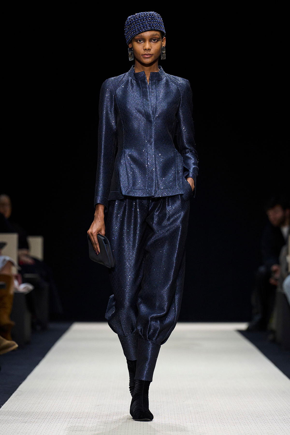 Female model walks a fashion runway in a sparkling navy blue suit with a matching sequined cap, holding a small clutch.