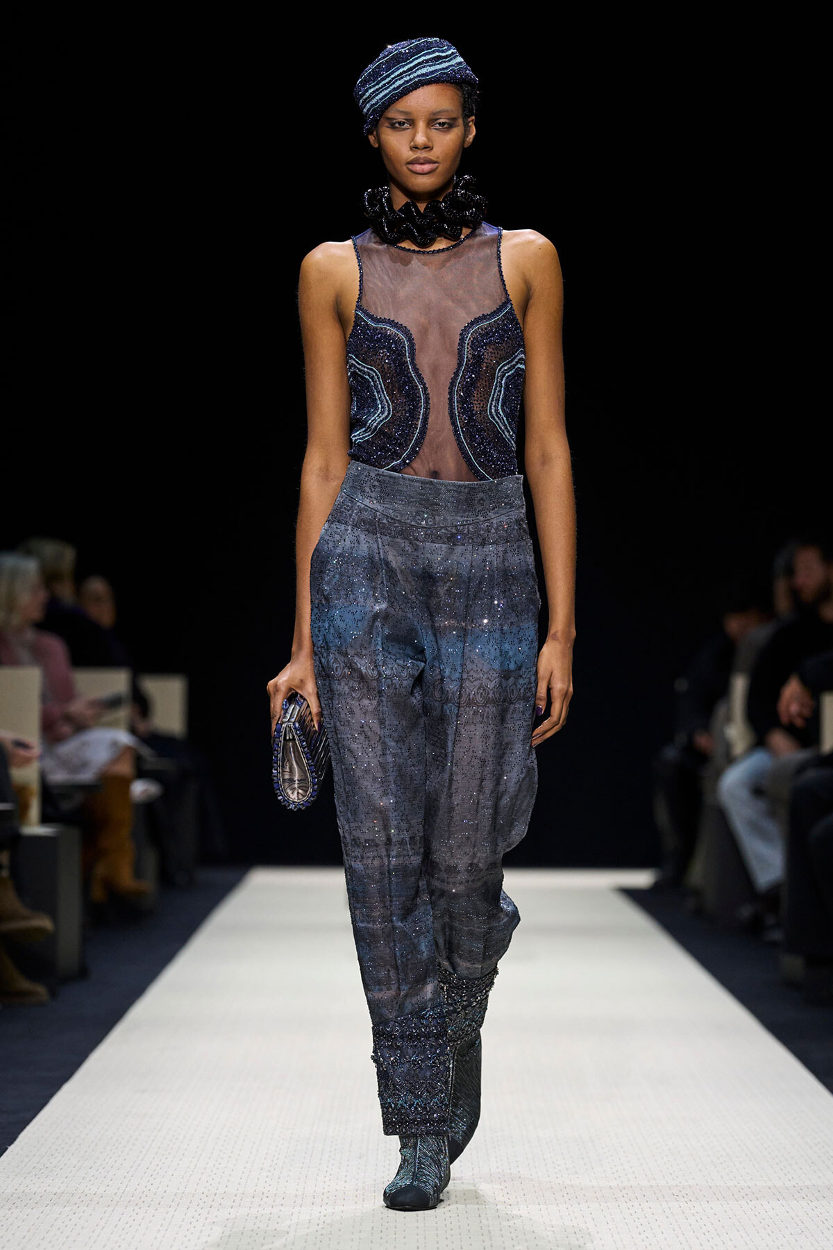 Runway model wearing a blue beaded sheer bodice with sparkling wide-leg pants and a matching blue turban, walking a white runway.