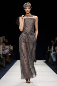 Model walks the runway in a shimmering gray, sheer, strapless gown with a wrapped waist and a beaded overlay, accessorized with a jeweled headwrap and layered necklace