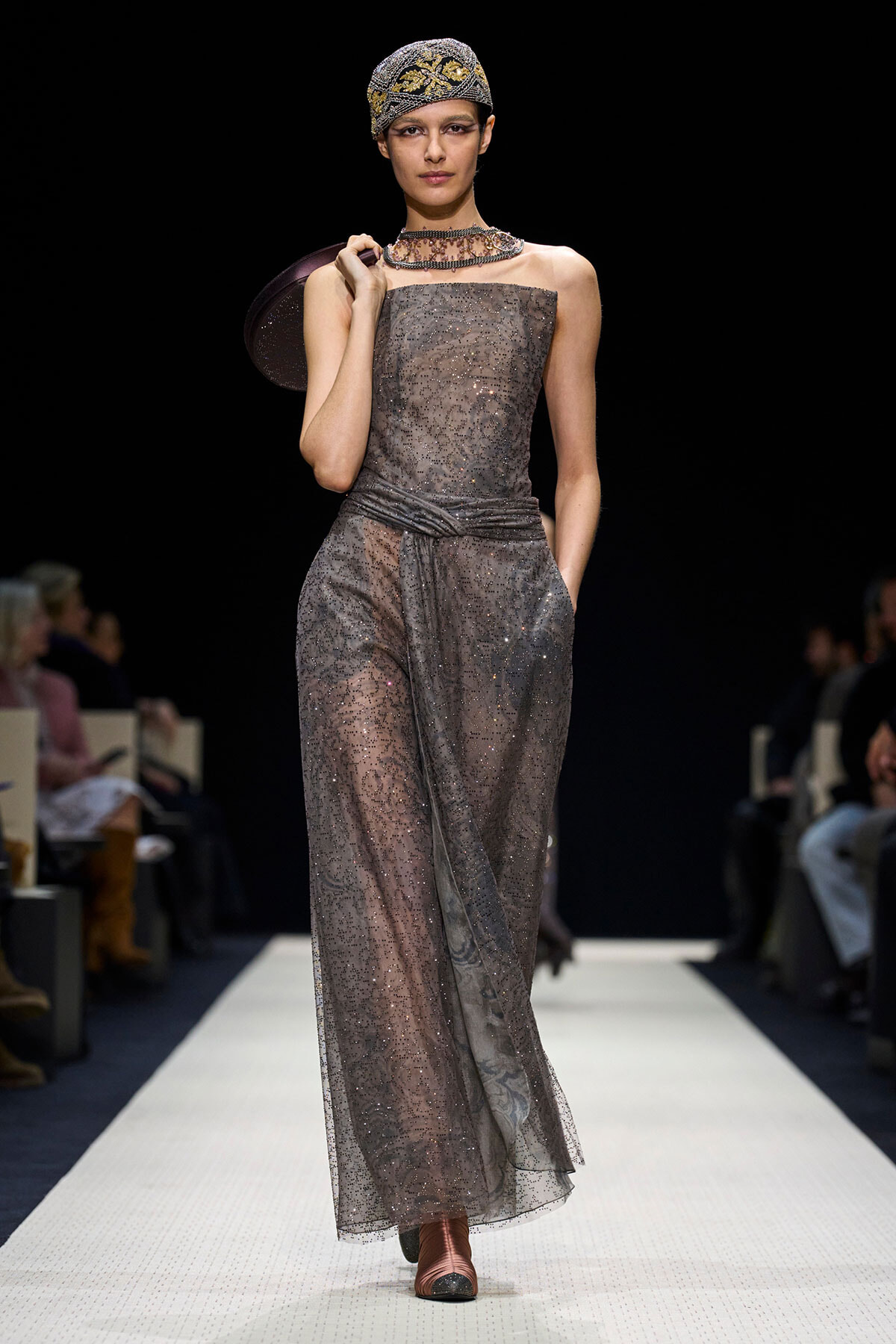 Model walks the runway in a shimmering gray, sheer, strapless gown with a wrapped waist and a beaded overlay, accessorized with a jeweled headwrap and layered necklace
