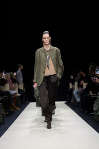 Model on a runway wearing an olive green blazer, beige top, and dark pants, holding a green bag while audience films on either side.