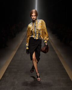 Model walks the runway in a gold and gray baroque-pattern blouse with leopard sleeves and a black draped skirt, carrying a brown handbag and wearing black gloves and heeled sandals