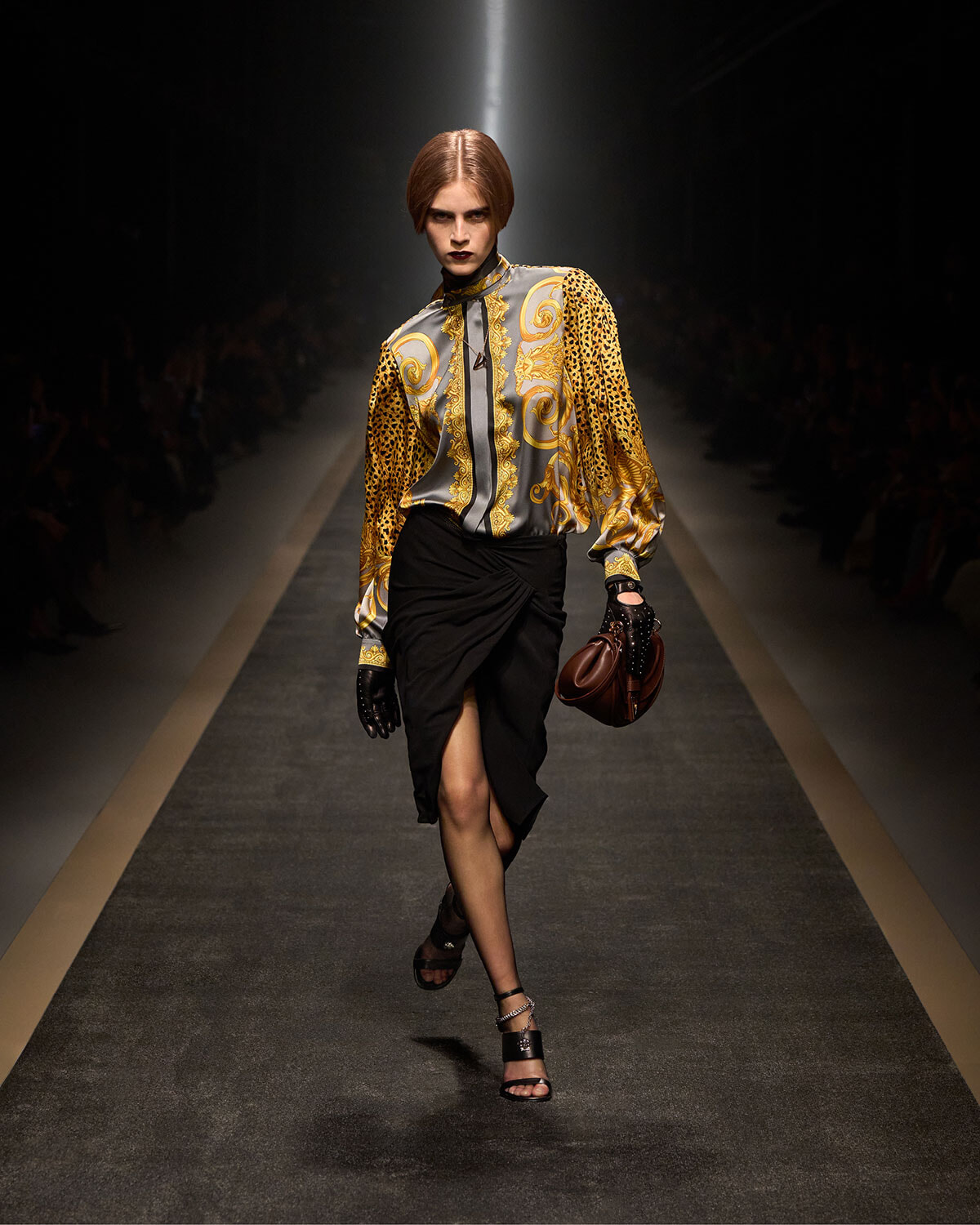 Model walks the runway in a gold and gray baroque-pattern blouse with leopard sleeves and a black draped skirt, carrying a brown handbag and wearing black gloves and heeled sandals
