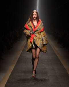 Model walks the runway in an oversized patterned puffer coat with red trim and leopard accents, spotlight behind.