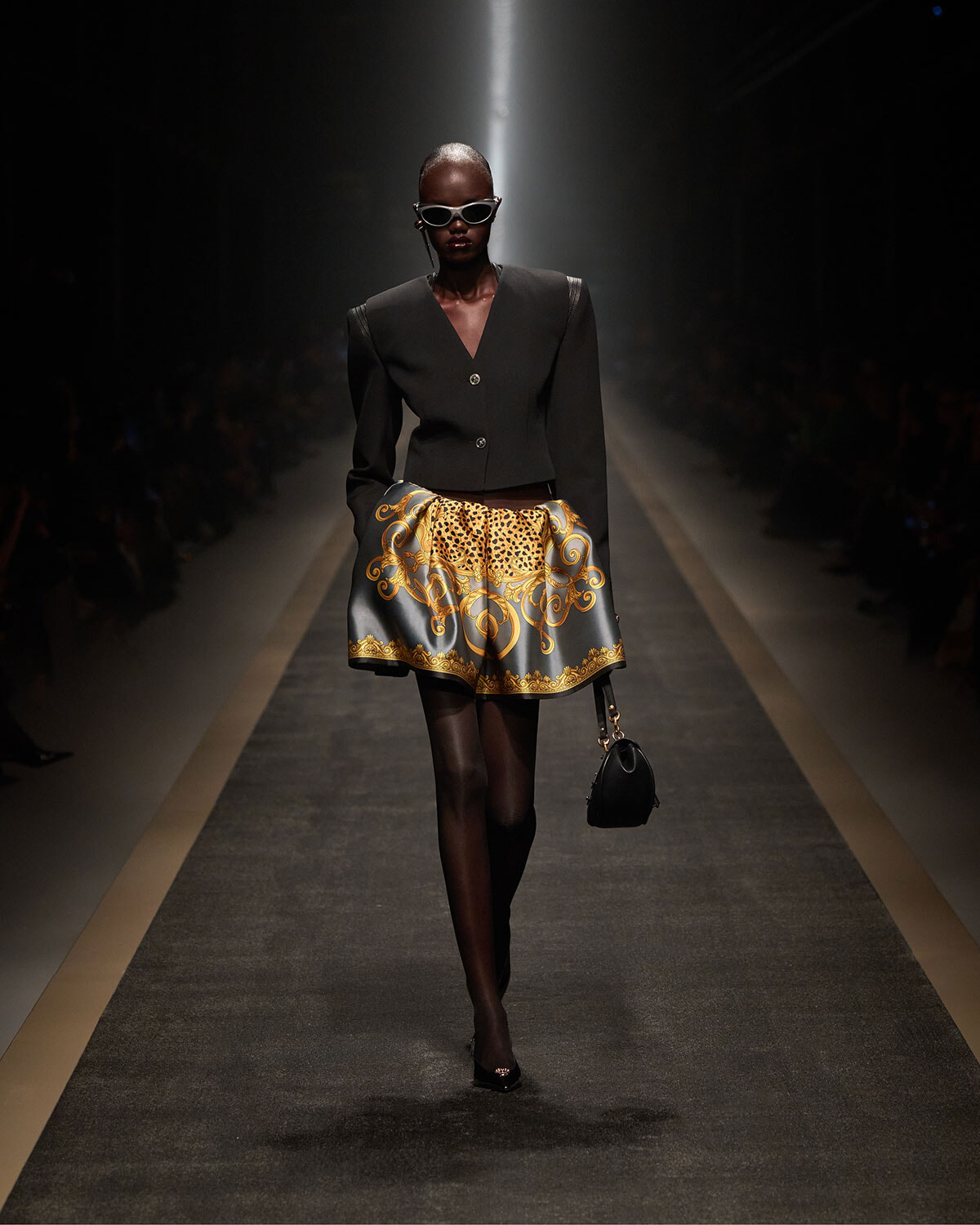 Runway model in a tailored black blazer and a voluminous gold-baroque print skirt, with sunglasses and a small black handbag on the catwalk.