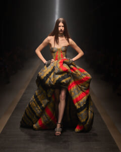 Model on a runway wearing a strapless, gold-black-red brocade ball gown with a voluminous skirt, hands on hips.