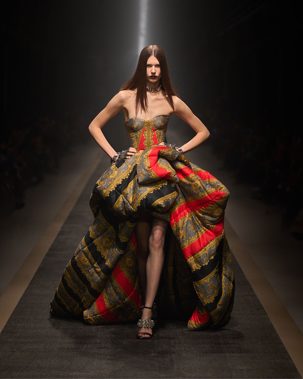 Model on a runway wearing a strapless, gold-black-red brocade ball gown with a voluminous skirt, hands on hips.