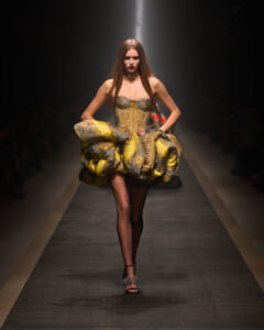 Runway look: a model wears a gold-and-blue brocade gown with a voluminous sculpted skirt, black tights, and spiked-heel sandals under a spotlight.