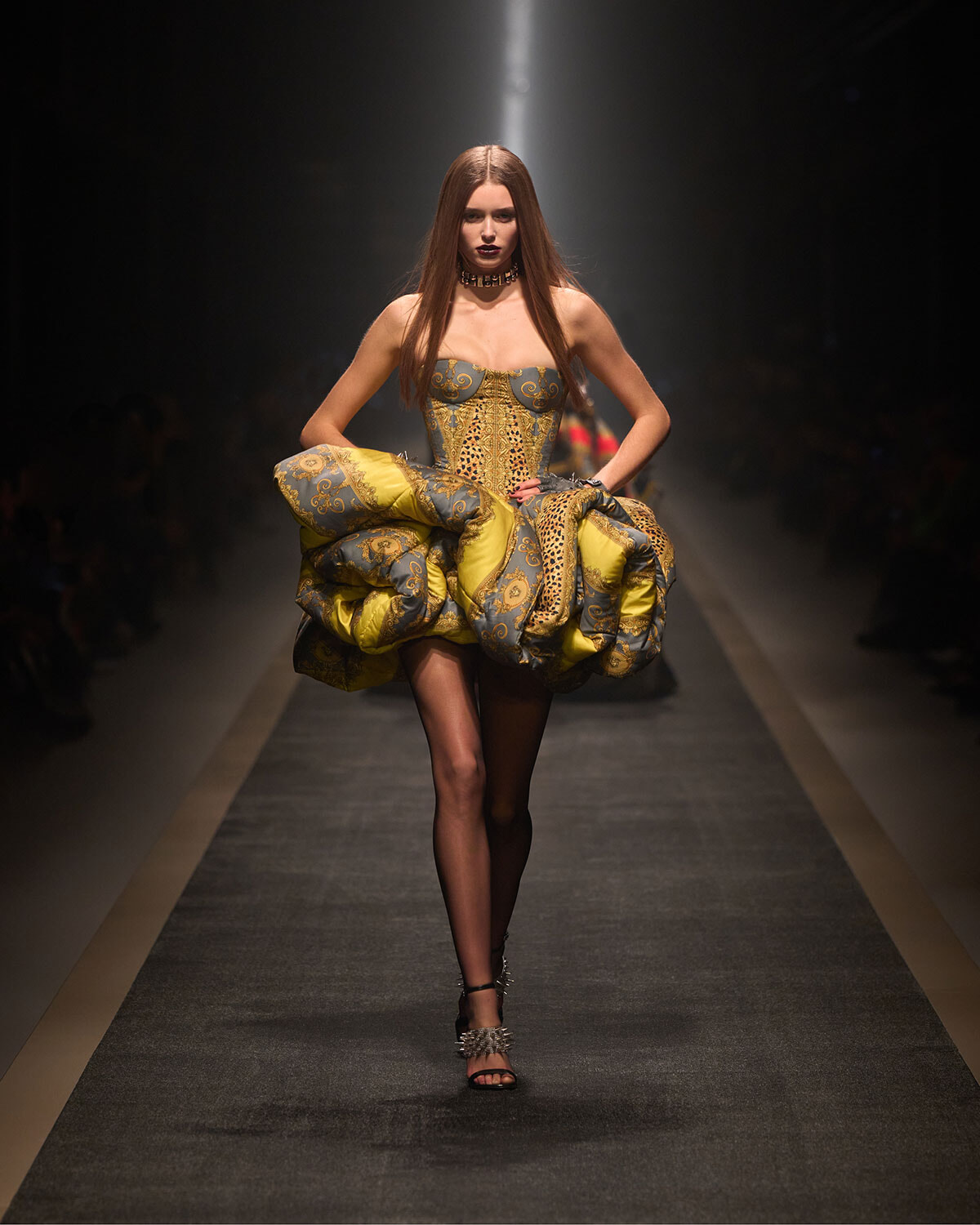Runway look: a model wears a gold-and-blue brocade gown with a voluminous sculpted skirt, black tights, and spiked-heel sandals under a spotlight.