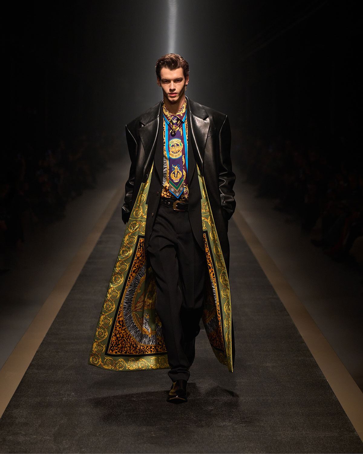 Male model strutting down a runway wearing a black leather coat with a green‑and‑gold patterned lining and a blue ornate scarf/tie.