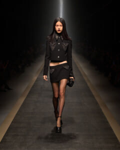 Model on a runway wearing a black cropped jacket with silver studs and a matching mini skirt with studded pockets.