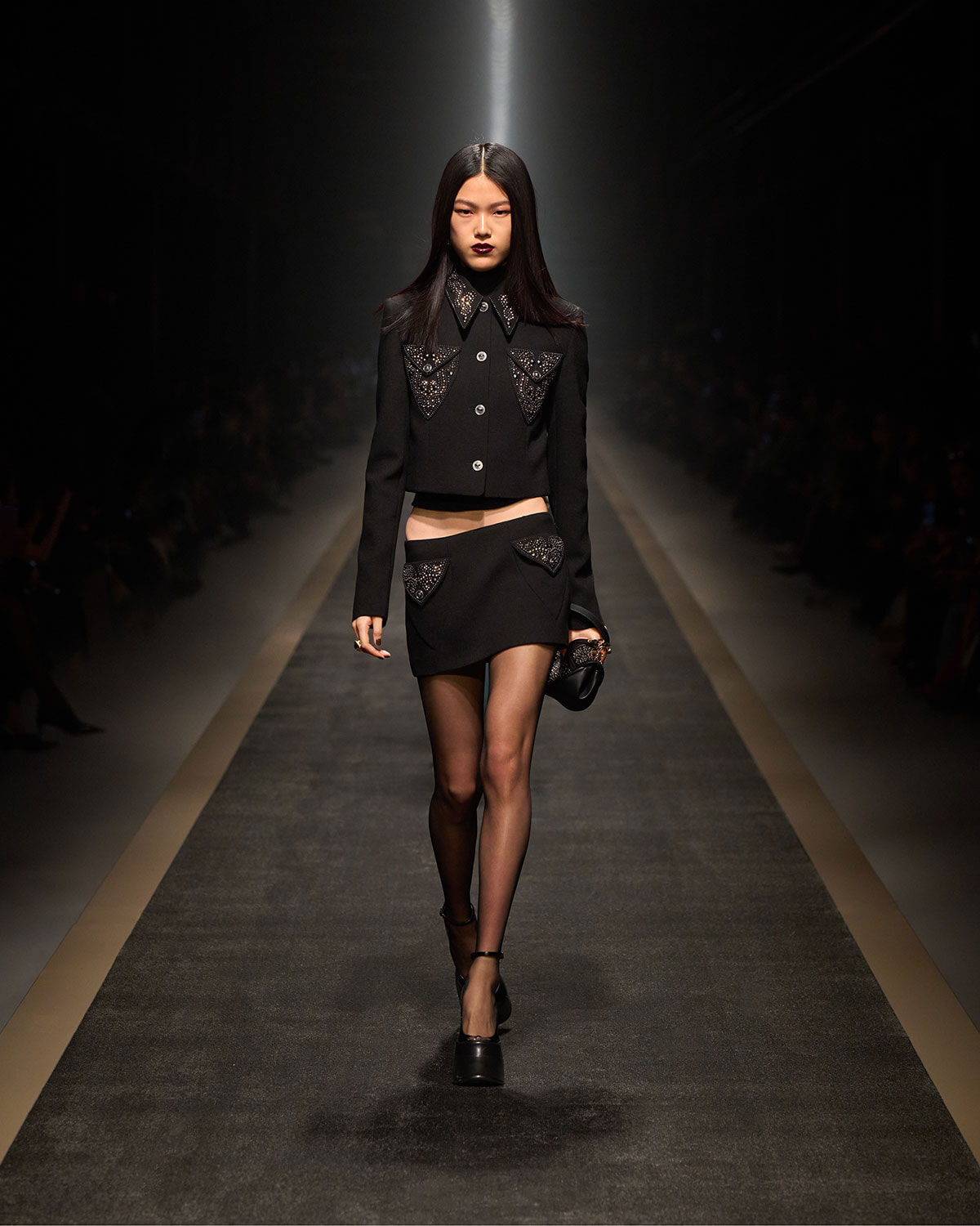 Model on a runway wearing a black cropped jacket with silver studs and a matching mini skirt with studded pockets.