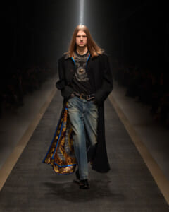 Male model with long hair walking a fashion runway in a black coat, graphic shirt, and distressed jeans, showcasing a colorful patterned lining peeking from the coat pocket side.
