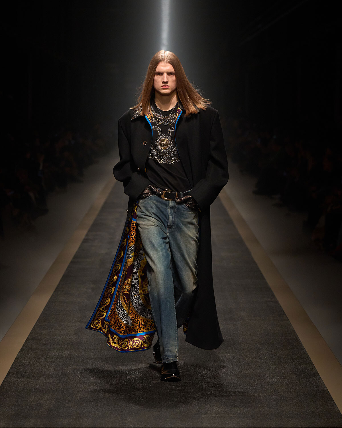 Male model with long hair walking a fashion runway in a black coat, graphic shirt, and distressed jeans, showcasing a colorful patterned lining peeking from the coat pocket side.