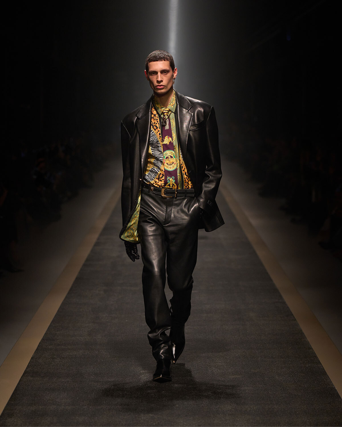 Male model struts a fashion runway wearing a black leather suit with a vibrant, patterned shirt and tie under dramatic spotlight.