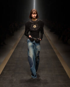 Model walking runway in a black embellished top and distressed jeans, wearing studded gloves under spotlight.