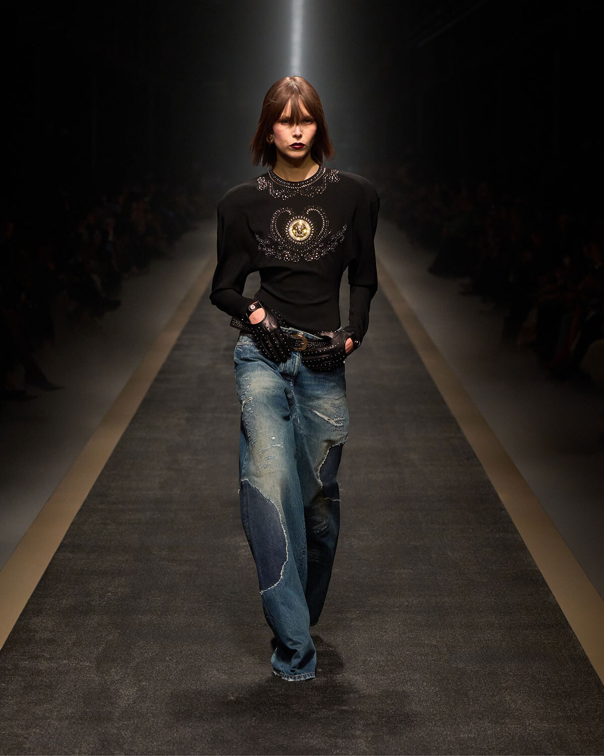Model walking runway in a black embellished top and distressed jeans, wearing studded gloves under spotlight.