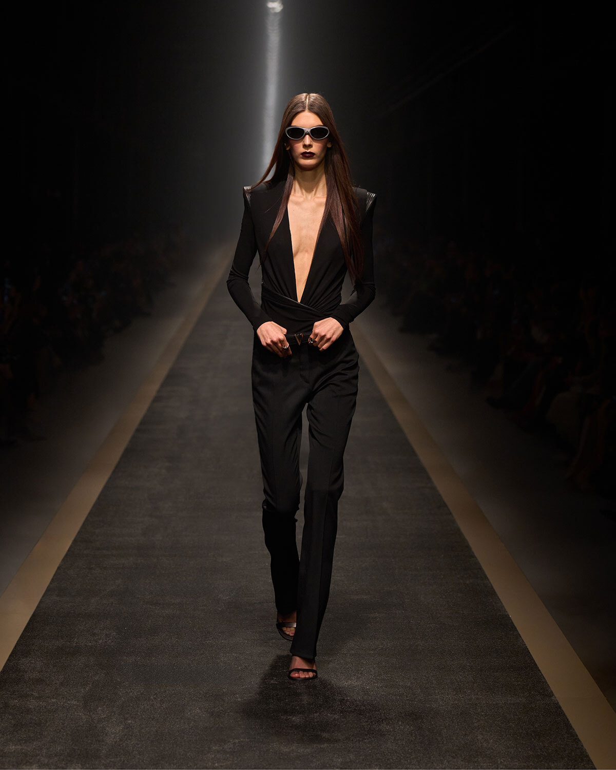 Model walking a dimly lit runway in a black plunging-neckline jumpsuit with shoulder pads and white sunglasses, spotlight behind her.