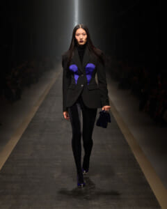 Female model walks runway wearing a black blazer with blue tonal panels, black turtleneck, tights, and matching blue handbag on her right arm.