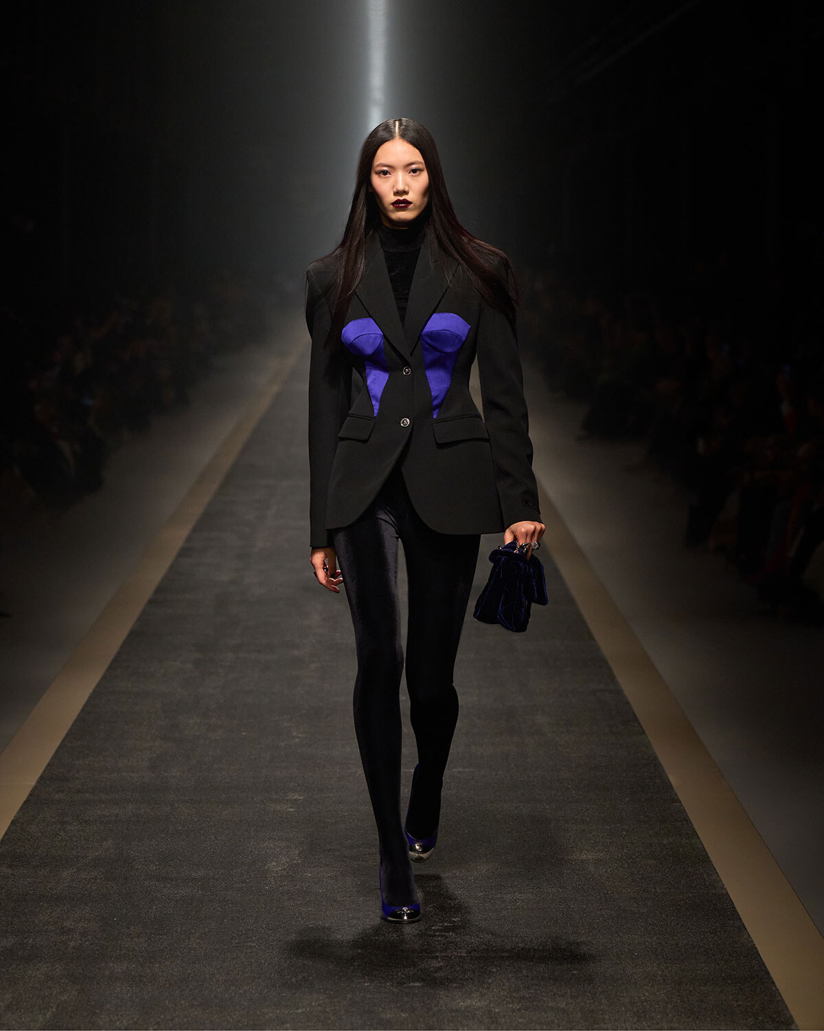 Female model walks runway wearing a black blazer with blue tonal panels, black turtleneck, tights, and matching blue handbag on her right arm.