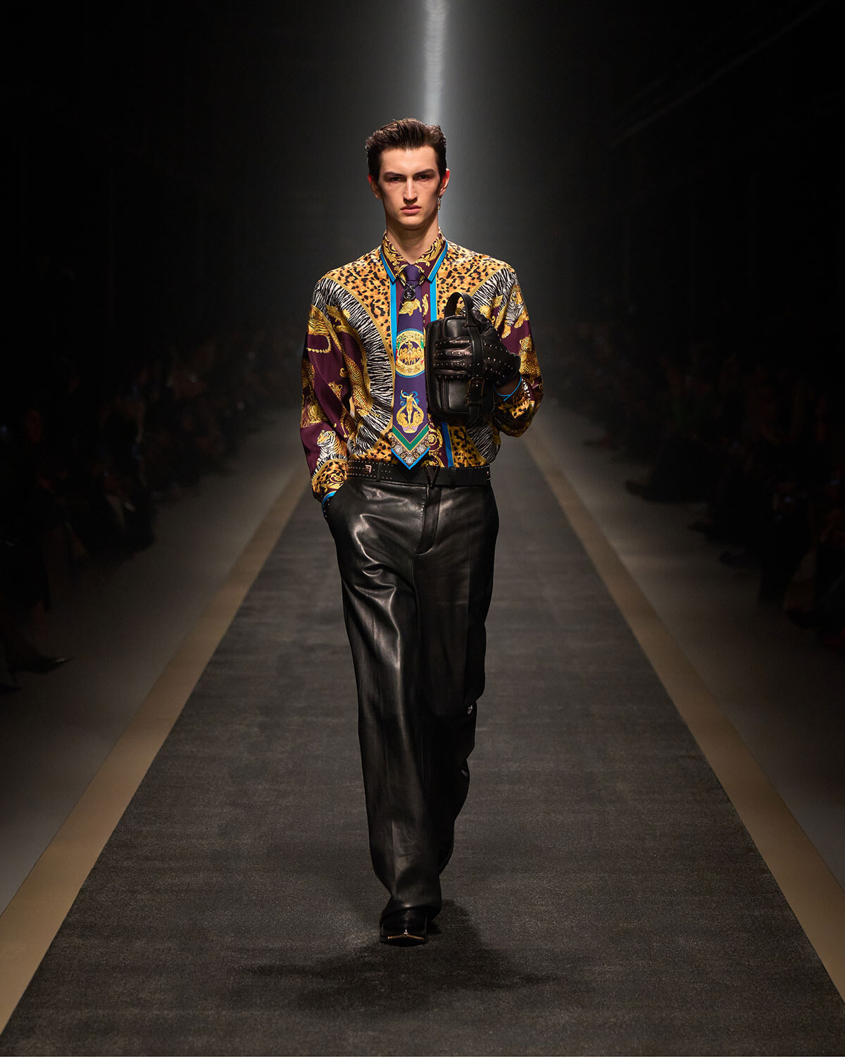 Male model on a fashion runway wearing a bold, multicolored patterned shirt, blue tie, black leather pants, and a black handheld handbag.