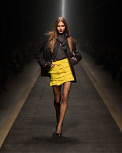 Model walking the runway in a black blazer over a textured black top, paired with a bright yellow layered mini skirt and black accessories under a spotlight.