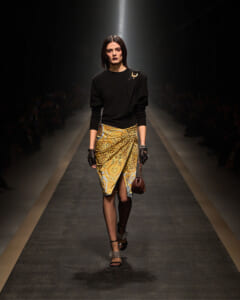 Model walks a runway in a black top and gold patterned wrap skirt with black gloves and a small brown handbag at a fashion show