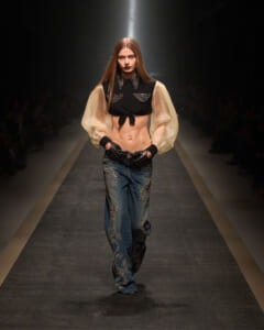 Model walking the runway in a cropped black top with sheer beige puff sleeves and embellished blue jeans with swirled embroidery.