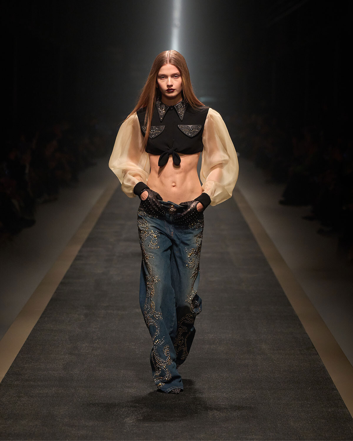 Model walking the runway in a cropped black top with sheer beige puff sleeves and embellished blue jeans with swirled embroidery.