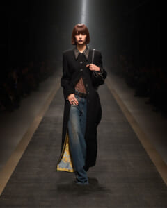 Model walks the runway in a knee-length black coat with a jeweled collar, wearing jeans and a sheer top, clutching a black handbag under spotlight.