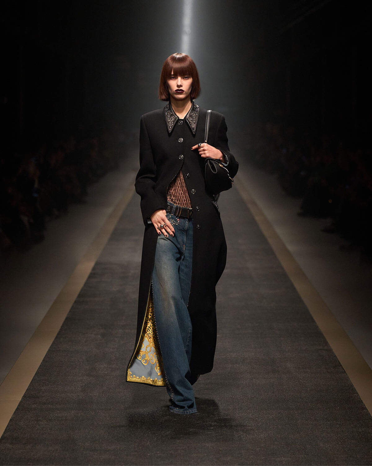Model walks the runway in a knee-length black coat with a jeweled collar, wearing jeans and a sheer top, clutching a black handbag under spotlight.