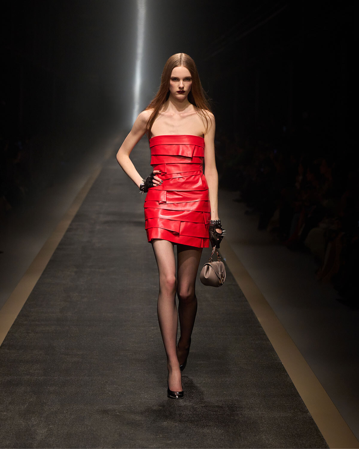 Model in a strapless red layered mini dress walks the runway, holding a small beige handbag.