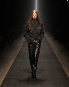 Model walking a runway in a black embellished shirt and shiny black leather pants, hands in pockets.