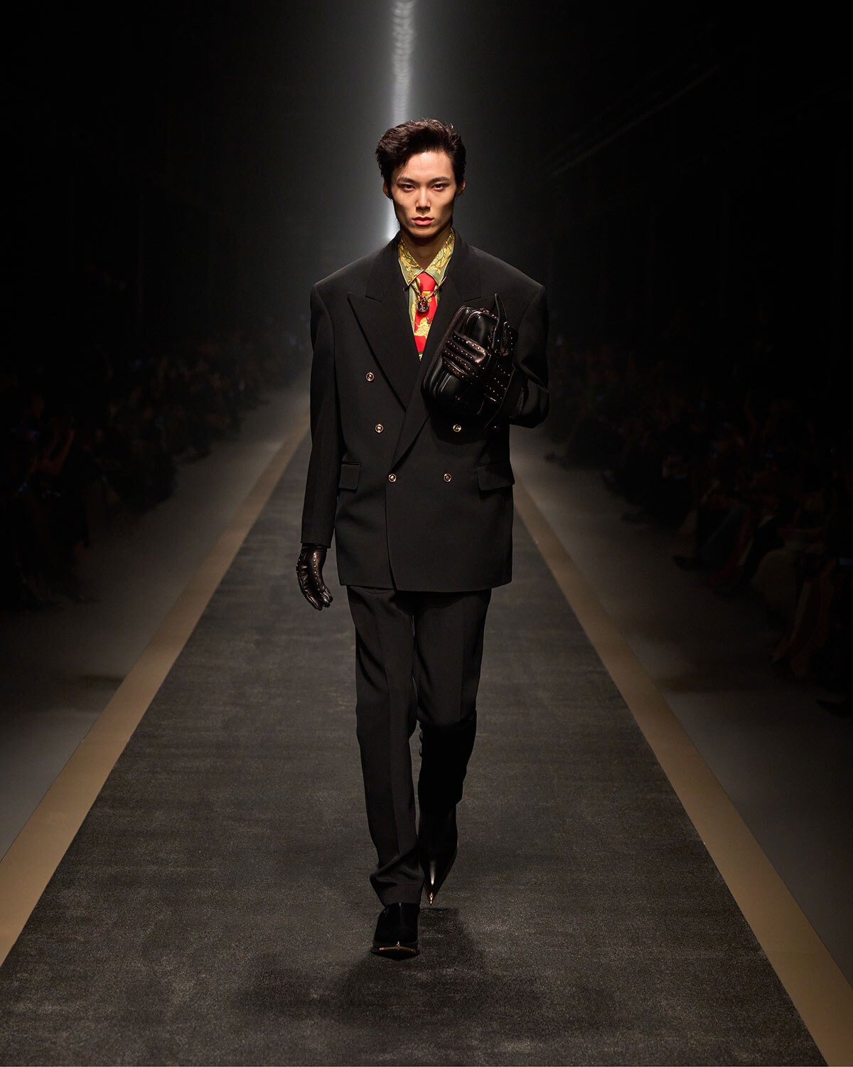 Male model walks a fashion runway in a black double-breasted suit, colorful patterned shirt, and black gloves held up under his arm.