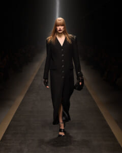 Model walks a runway in a long black buttoned coat with slit, black gloves, and strappy heels under spotlight.
