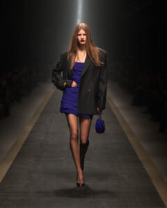 Model walking a fashion runway in a black pinstripe blazer over a cobalt blue tiered mini dress, carrying a small blue handbag and wearing black heels.