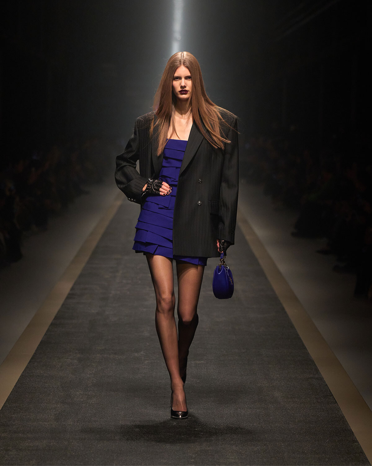 Model walking a fashion runway in a black pinstripe blazer over a cobalt blue tiered mini dress, carrying a small blue handbag and wearing black heels.