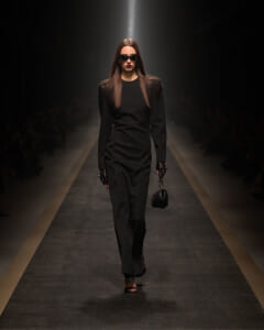 Model walks a dimly lit runway in a sleek black long-sleeve sheath dress, wearing sunglasses and carrying a small black handbag with a chain strap.