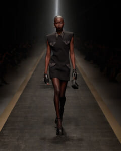 Female model walks down a dim runway in a black sleeveless shift dress with studded pocket details, gloves, and a small black handbag.