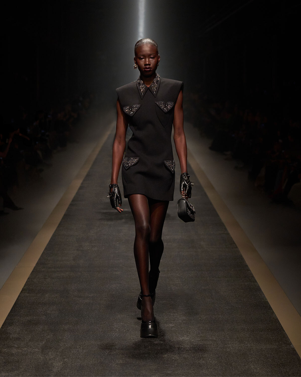 Female model walks down a dim runway in a black sleeveless shift dress with studded pocket details, gloves, and a small black handbag.