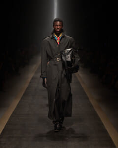 Male model walking a fashion runway in a long black trench coat with a belt, black leather gloves, and a large black bag slung over the shoulder/armlight.