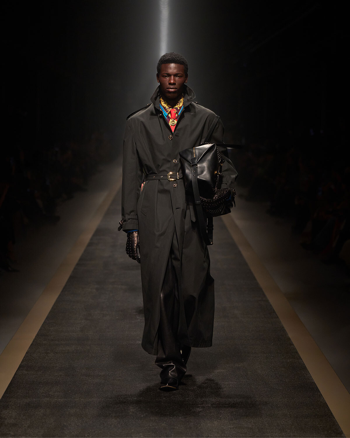 Male model walking a fashion runway in a long black trench coat with a belt, black leather gloves, and a large black bag slung over the shoulder/armlight.