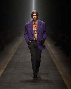 Model walking a runway in a purple blazer over a colorful patterned shirt, black trousers, and black gloves.