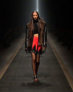 Model walking a fashion runway in a leopard-print and black leather jacket, black crop top, and a jagged red-and-black mini skirt.