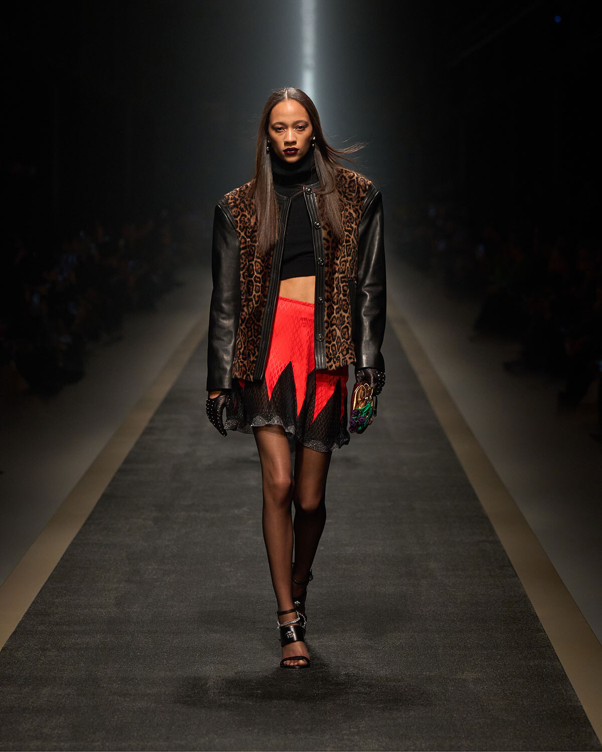 Model walking a fashion runway in a leopard-print and black leather jacket, black crop top, and a jagged red-and-black mini skirt.