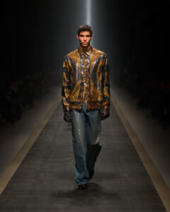 Male model walking a fashion runway in a gold-leopard and metallic jacket with intricate swirls, dark gloves, and wide-leg jeans.