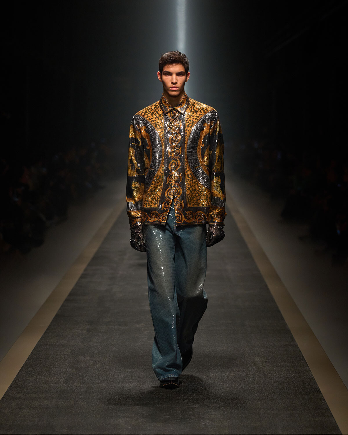 Male model walking a fashion runway in a gold-leopard and metallic jacket with intricate swirls, dark gloves, and wide-leg jeans.